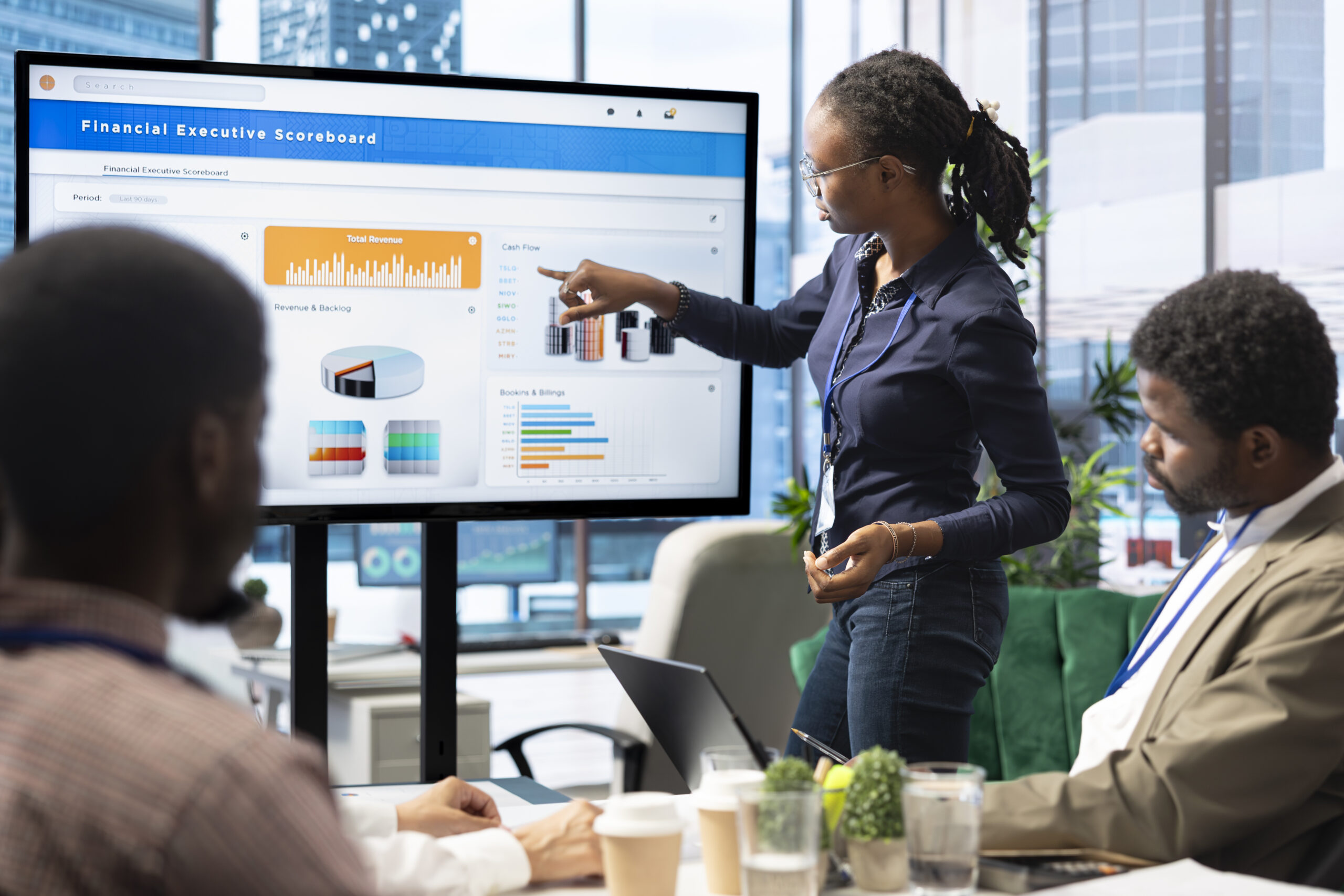 Uma profissional negra apresenta gráficos e indicadores financeiros em uma tela grande durante uma reunião corporativa. Ela está em pé, apontando para o painel intitulado “Financial Executive Scoreboard”, que exibe gráficos de barras, pizza e colunas coloridas sobre receitas, fluxo de caixa e desempenho de negócios. Dois colegas negros observam atentamente a apresentação, sentados à mesa com notebooks, copos d’água e plantas decorativas. O ambiente é moderno, com janelas amplas que mostram prédios corporativos ao fundo e luz natural iluminando o espaço.